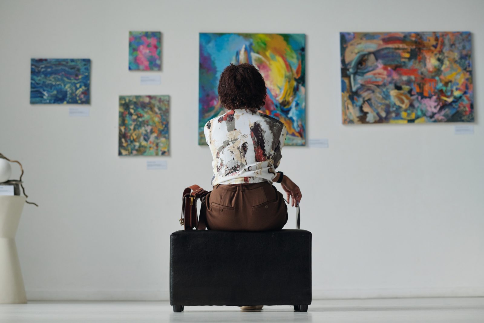 A person with their back towards the camera sits observing five paintings on a gallery wall.