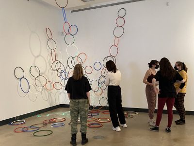 5 gallery viewers in front of art installation of painting and large coloured hoops hanging from ceiling