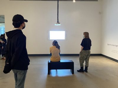 3 gallery attendees viewing projection on a wall, one sitting on a bench