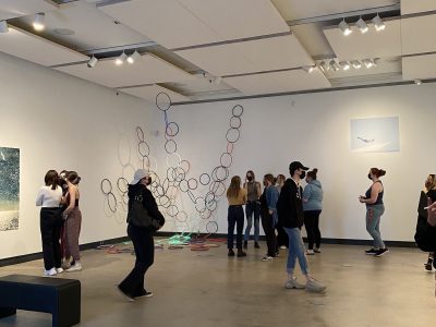 many gallery attendees in front of art instalation of painting and large coloured hoops hanging from ceiling