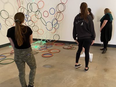 4 gallery viewers in front of art instalation of painting and large coloured hoops hanging from ceiling