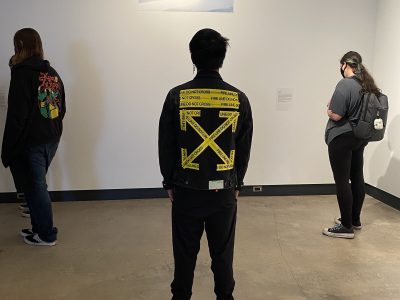 3 gallery attendees looking at abstract painting, on male has leather jacket with yellow arrows painted on back