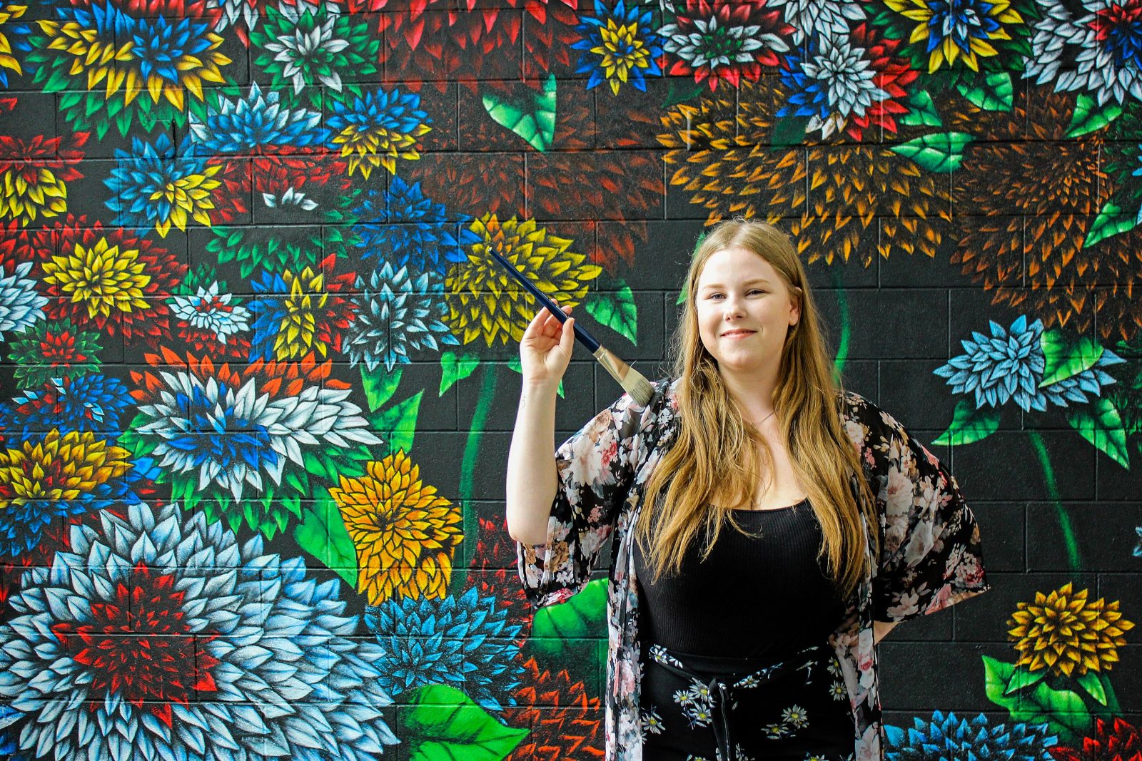 Fourth-year Visual Arts student Chardon Trimble-Kirk holding paintbrush in front of completed mural