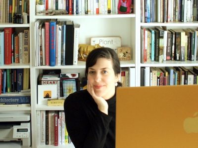 A woman wearing a black shirt sits at a desk looking at the camera from behind a computer screen with a bookshelf with colourfu; books in the background.