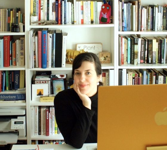 A woman wearing a black shirt sits at a desk looking at the camera from behind a computer screen with a bookshelf with colourfu; books in the background.