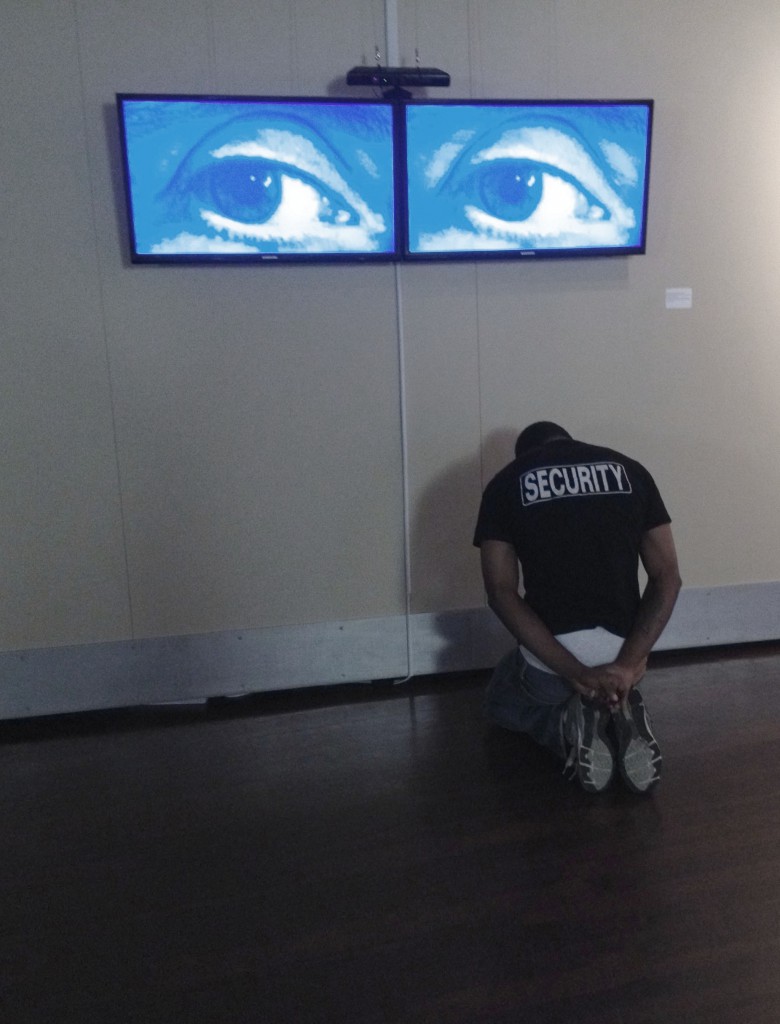 Szoke Lexie Mountain, securty officer on knees facing wall with 2 tv screens displaying human eyes