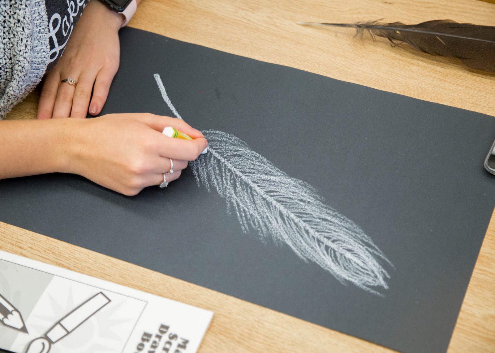 woman's hands drawing a white chalk feather on black paper