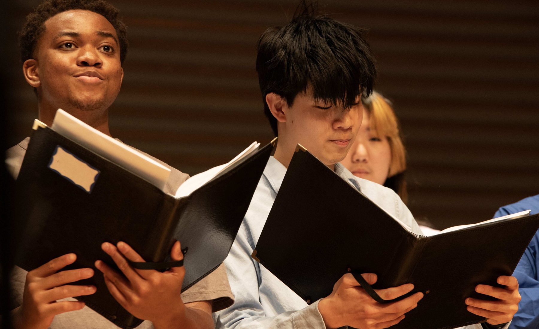 Three students holding black music folders are poised to sing. 