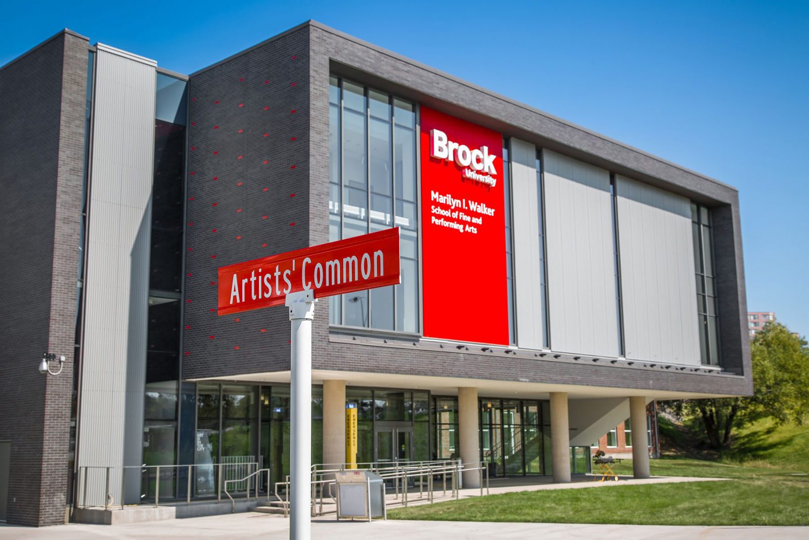 A grey and red building sits against a clear, blue sky with green grass surrounding it. The building has white text on it on a red background reading ‘Marilyn I. Walker School of Fine and Performing Arts, Brock University.’ A street sign reading ‘Artists’ Common’ sits in the foreground.