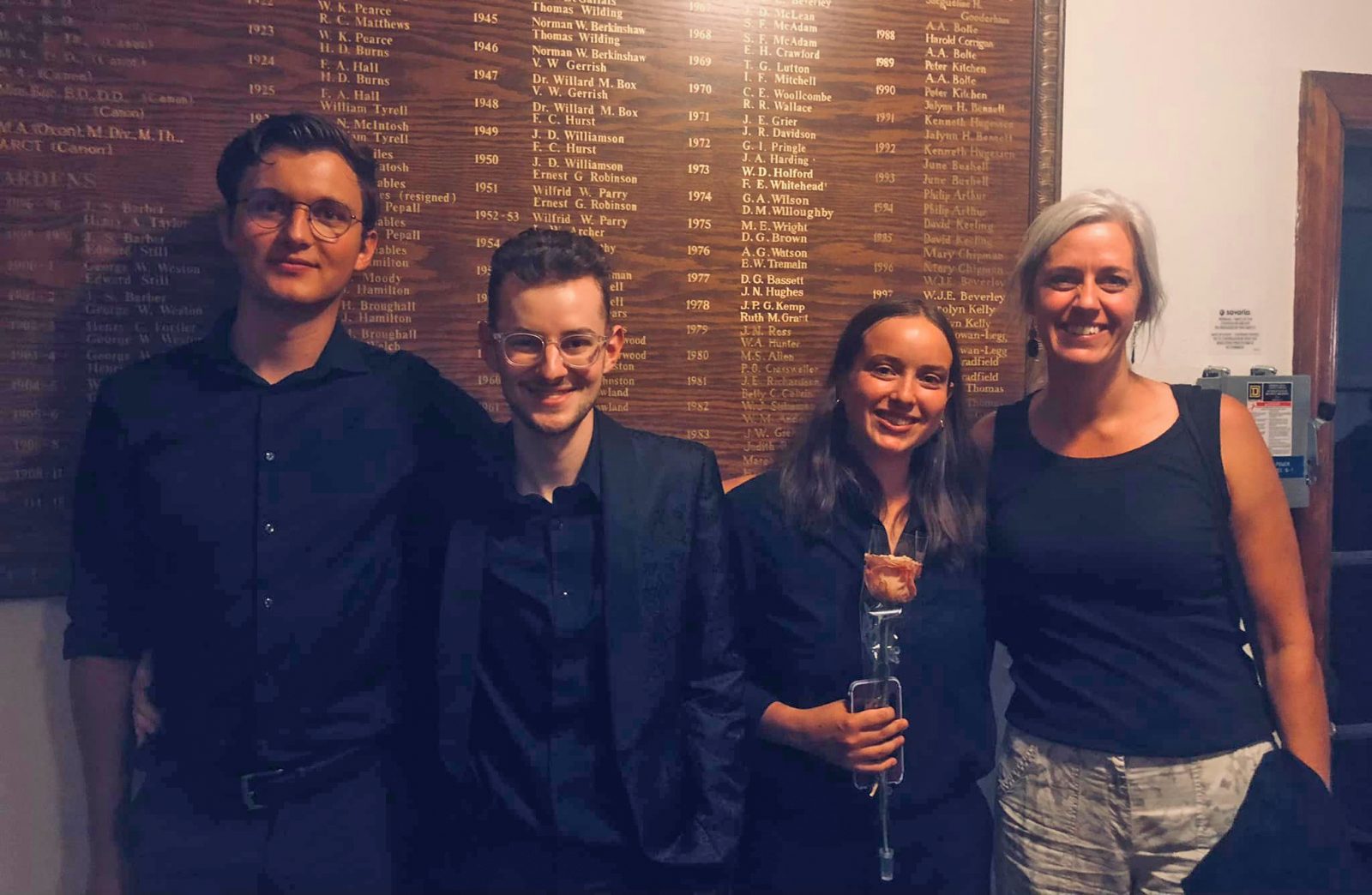 An image of a group of four individuals: two men on the left and two women on the right, all wearing black semi-formal wear. There is a wooden wall behind them with names of recognized individuals and years