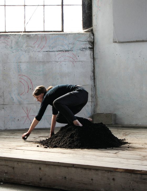 Artist on top of dirt pile drawing with dirt on floor of interior of industrial building