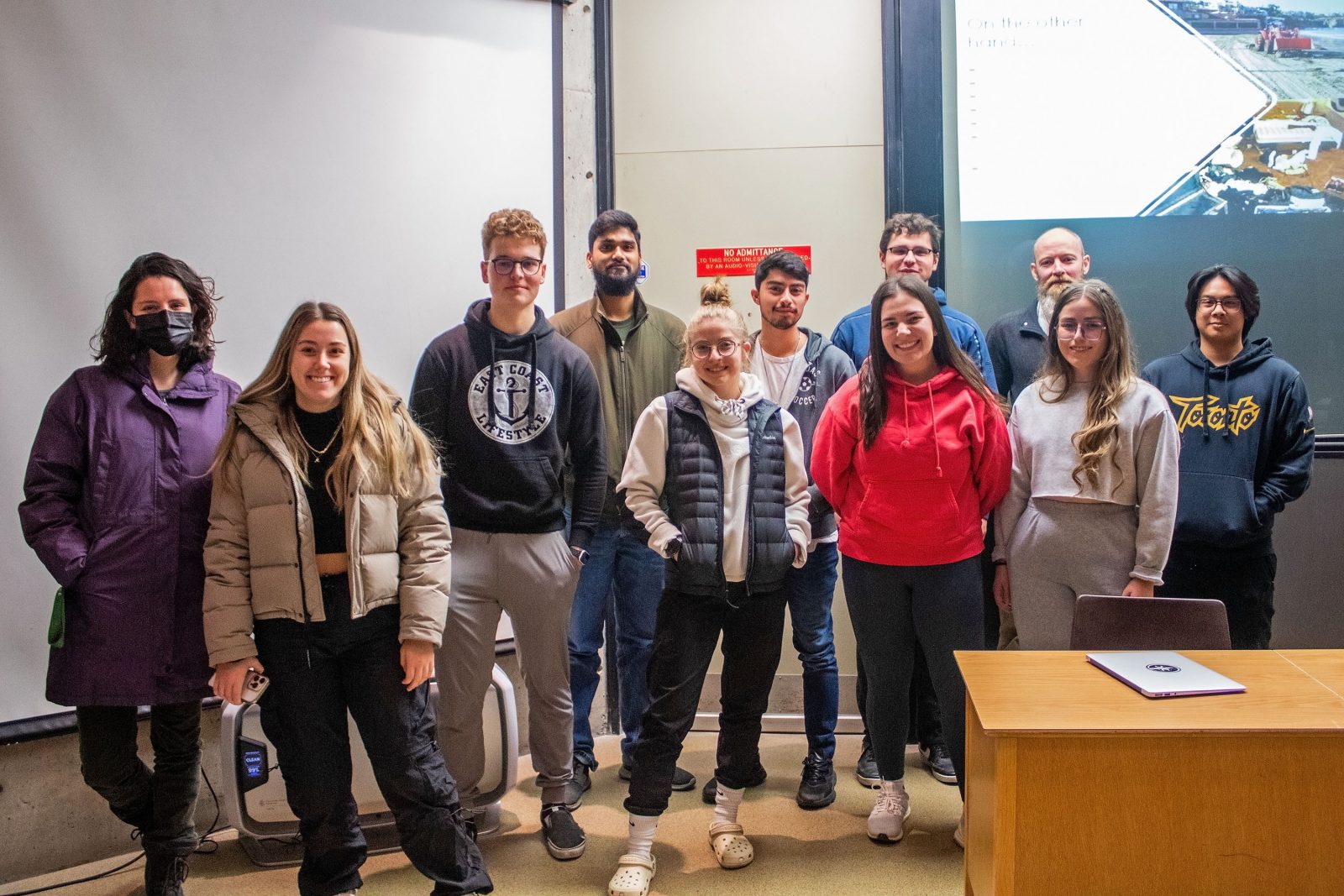A group of students stand at the front of a lecture hall in front of two large projector screens.