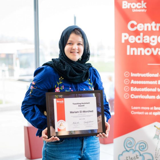 Mariam El-Morched smiles holding her TA Award in front of the CPI banner