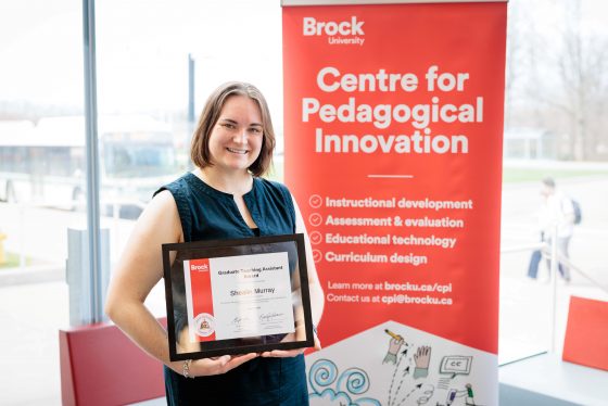 Shealin Murray smiles holding her Graduate TA Award in front of the CPI Banner