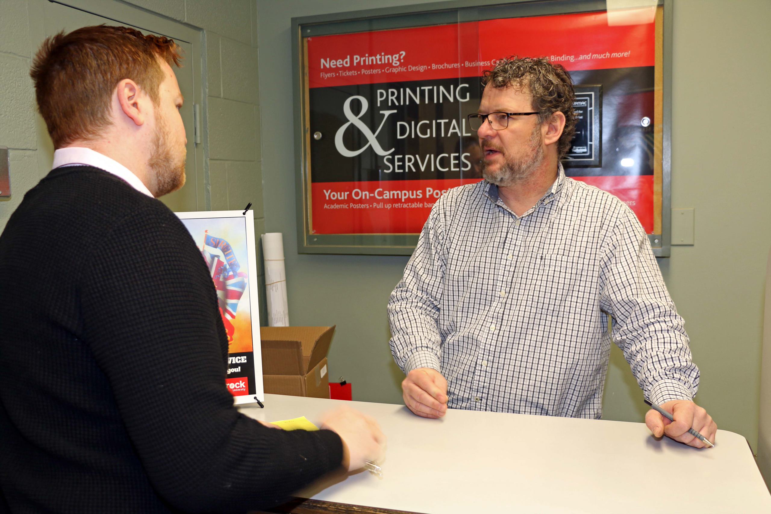 A medium shot shows two men at a counter; the man in the foreground is wearing a black sweater and the man behind the counter is in a checkered shirt. The background shows a sign that reads "Printing & Digital Services."