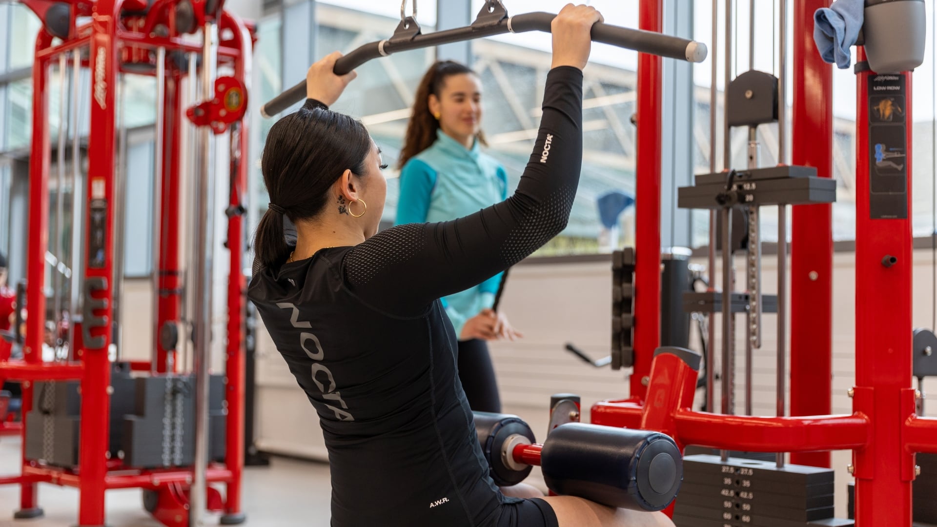Asian and Spanish female students working out on gym equipment smiling 
