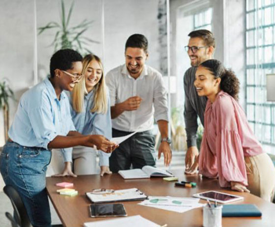 A diverse group of colleagues gathers around a table, smiling and pointing at documents. This image represents the training aspect of the Step-by-Step Parenting Program.