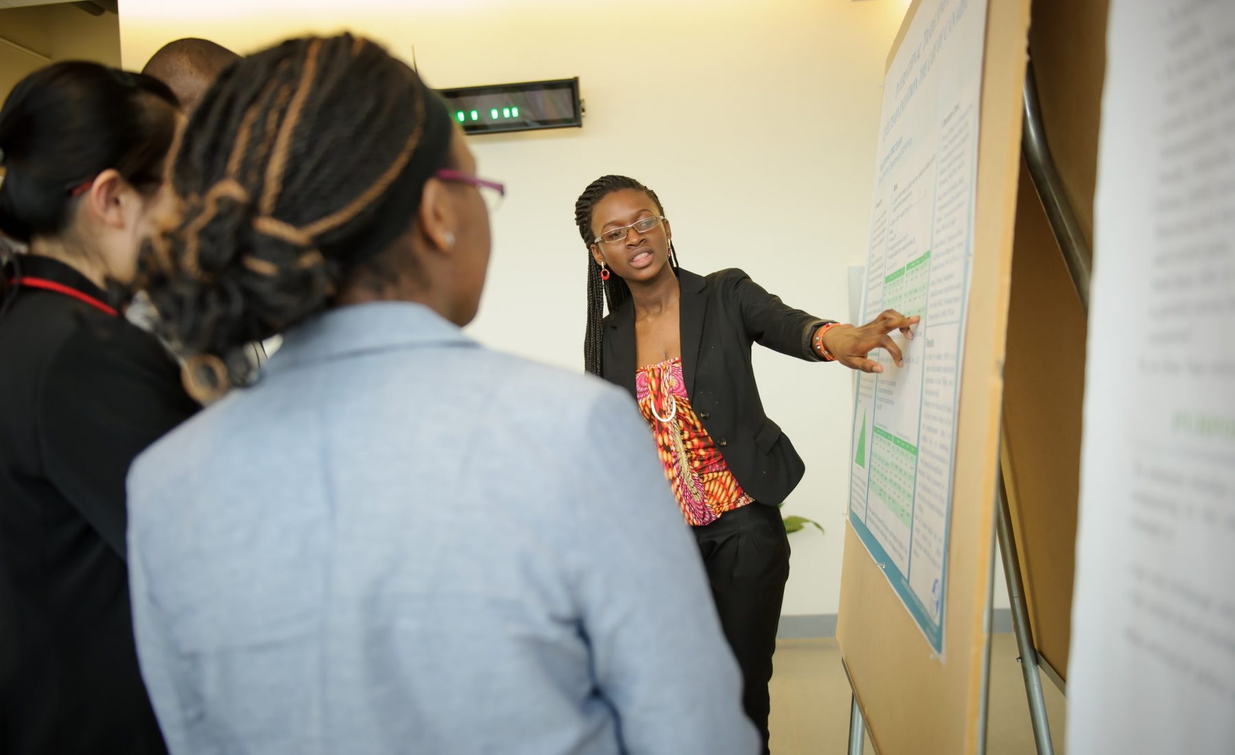 Person presenting to a small group and pointing at a poster that has text and graphs. 