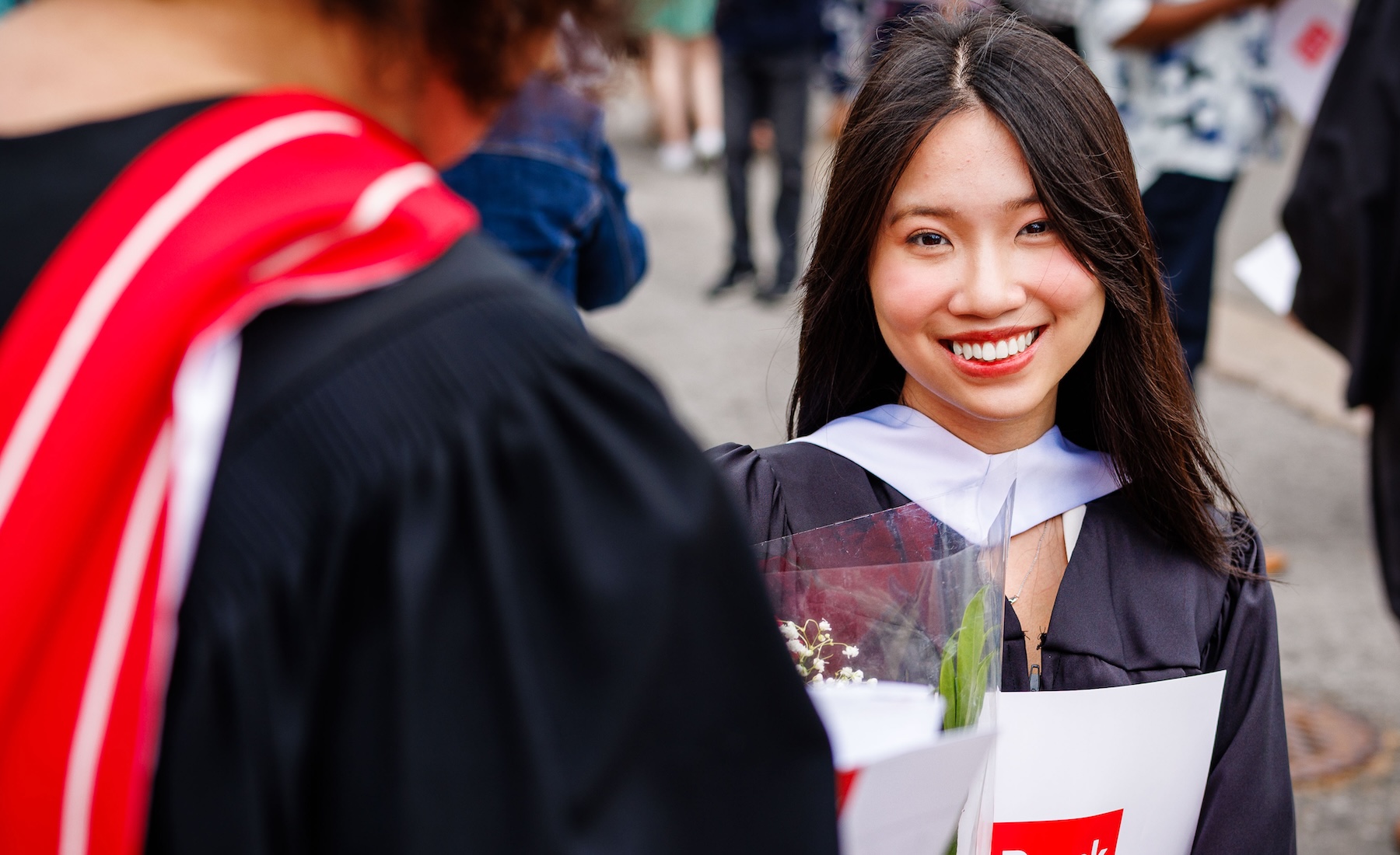 A graduate in a Convocation gown smiles while holding flowers and a Brock University branded folder.
