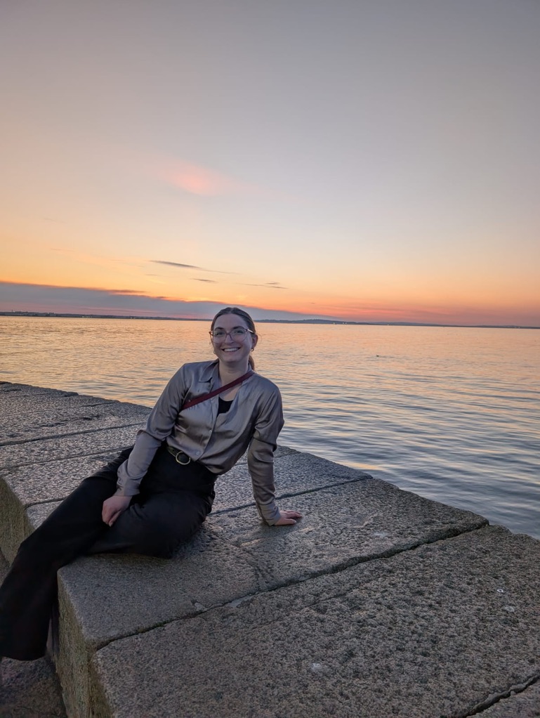 Elizabeth Al-Jbouri sitting on a stone wall with a sunset over the water in the background.