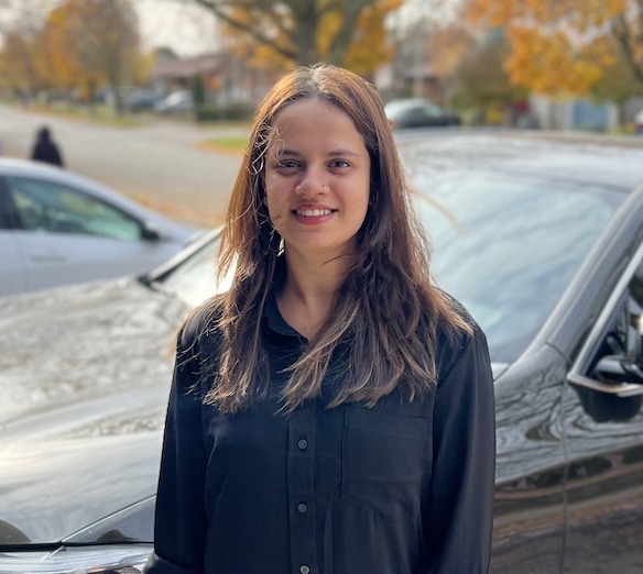 A person standing in front of a car in a parking lot with trees in the background.