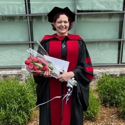 Lisa Whittingham wearing a graduation gown and cap while holding a bouquet of red roses and posing for a photo outside on Brock University's main campus.