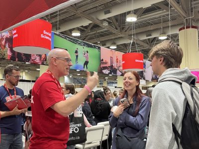 A man in a Red Brock University shirt gestures as he speaks to a gathering of students, professors and parents.