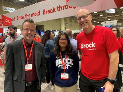 Two men, one in a red Brock University t-shirt and the other in a suit jacket with a red Brock lanyard, pose for a photo with a current student between them. The student is wearing a Brock University sweatshirt.
