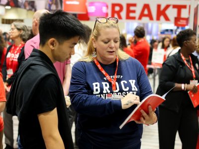 A woman in a blue Brock University shirt holds a booklet and is explaining something to a prospective student.