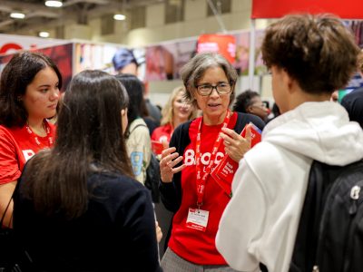 A professor in a red Brock University shirt is gesturing as they speak to a gathering of prospective students