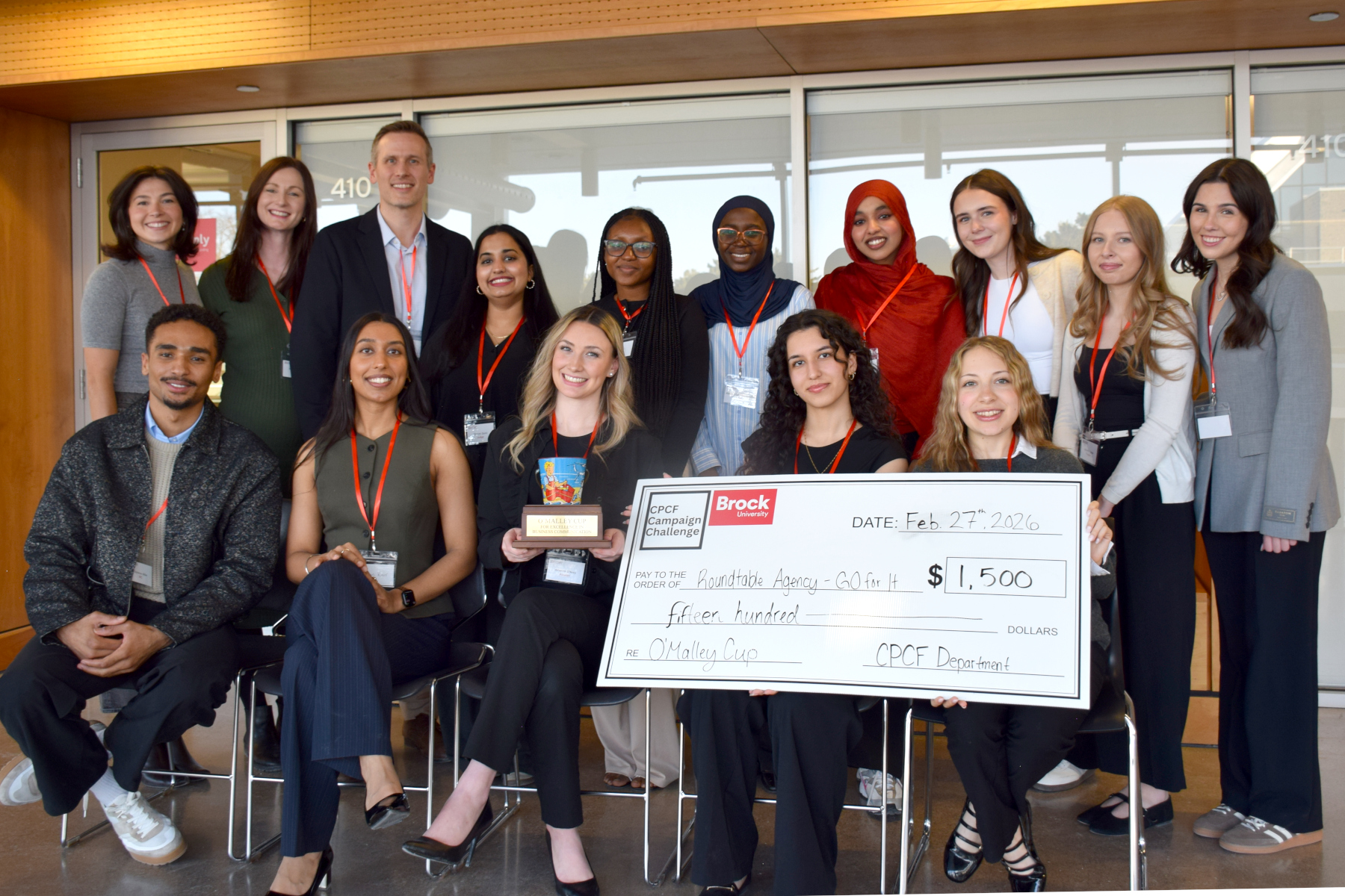 A group of 15 people stand together and pose for a photo. a few of the people in the front of the photo are holding a giant check and a trophy.