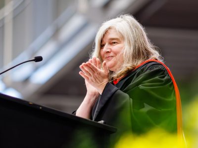 A professor in a convocation gown stands behind a podium clapping.