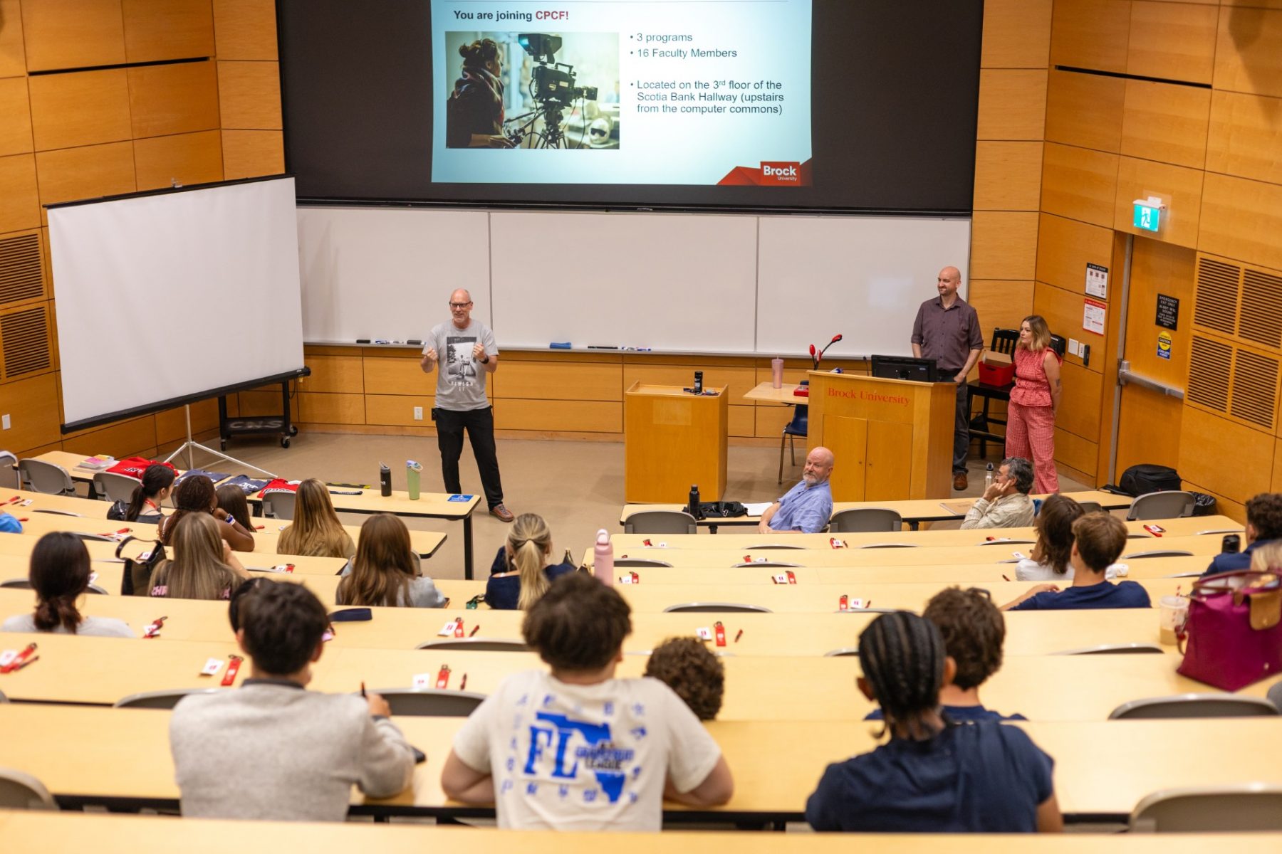 Faculty and staff are gathered at the front of a lecture hall with students watching them from the seats.
