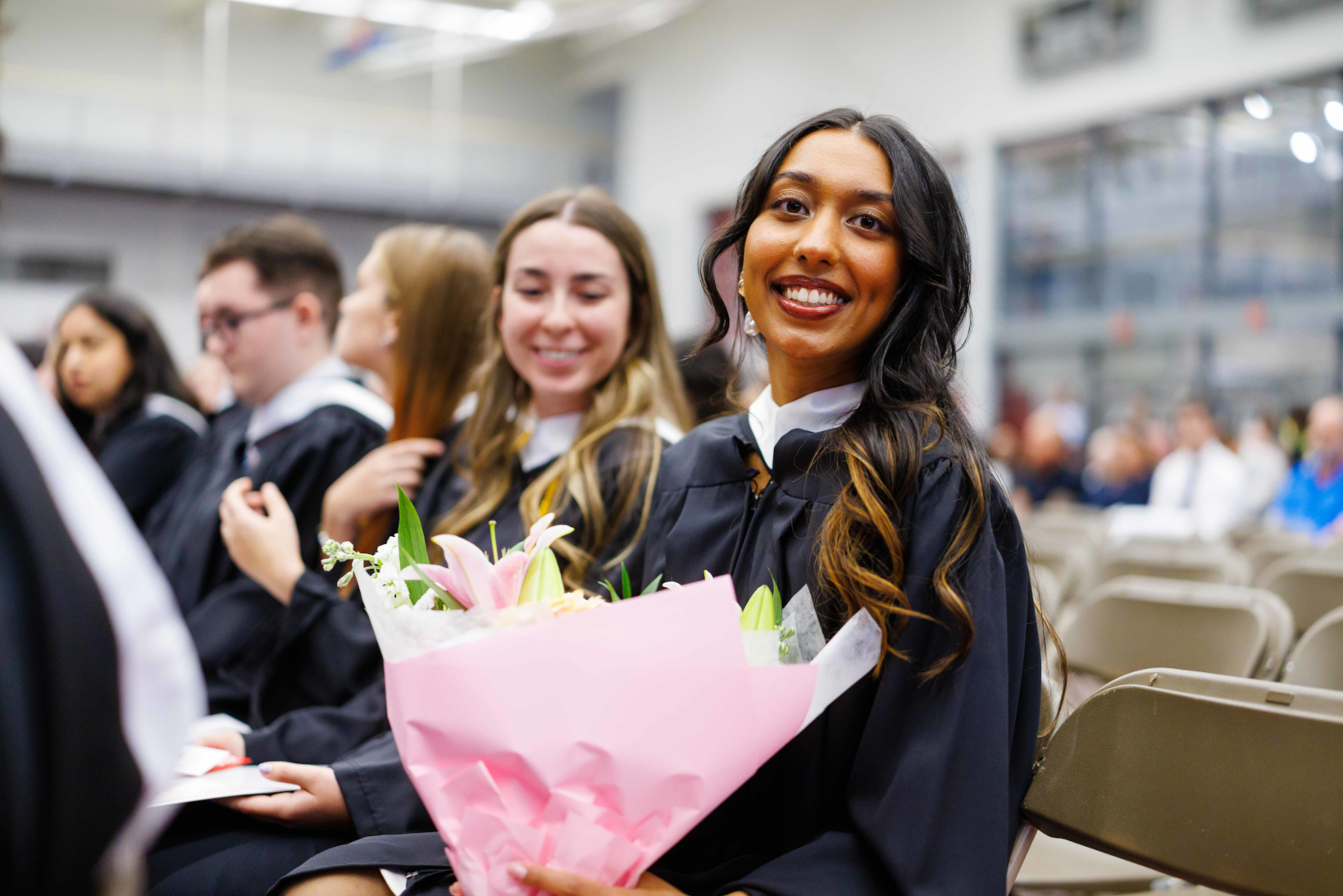 A student holding flowers and wearing a graduation gown smiles for a photo. They are surrounded by other students in gowns.