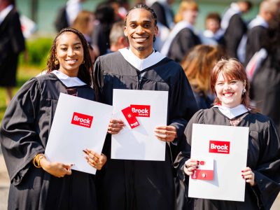 Three people in graduation gowns standing together outside and holding up their degrees.