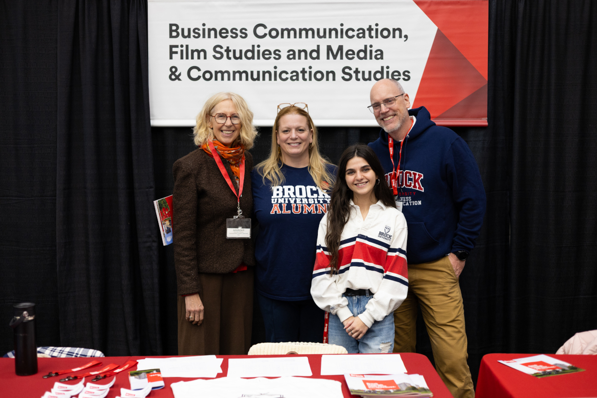 four people, some in Brock University clothing, stand behind a table and pose for a photo. The sign behind them identifies their department programs as Buisnes Communication, Media and Communication and Film Studies