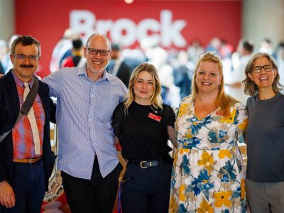 Five people posing for a photo with their arms around each other in Brock University's Rankin Family Pavilion Atrium.