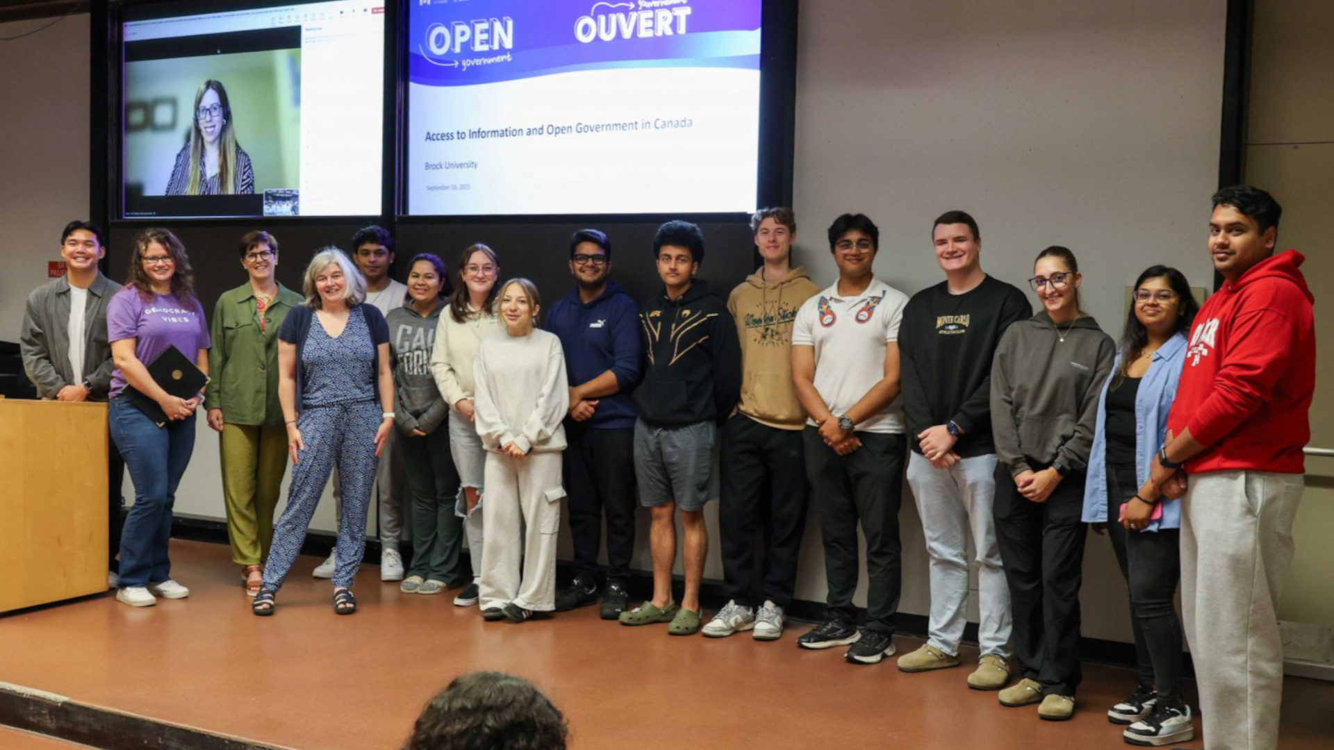 A lineup of the professor, their students and two guest speaks at a podium. They are standing at the front of an auditorium. Behind them is a screen featuring the face of the third presenter. 
