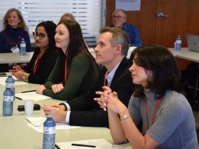 A panel of four professional looking people sitting at a desk in a conference room setting.