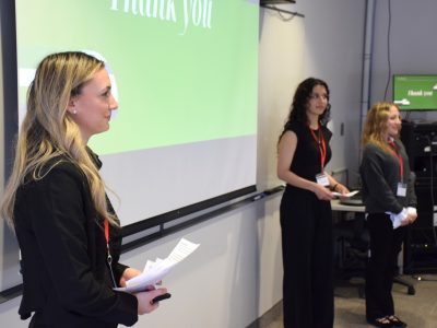 three students, professionally dressed and wearing name tags give a presentation to an unseen audience. They are standing in front of a projected screen.
