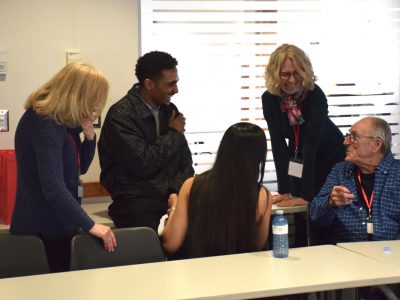 A group of people both young adults and adult professionals are speaking and laughing together in a conference room setting.