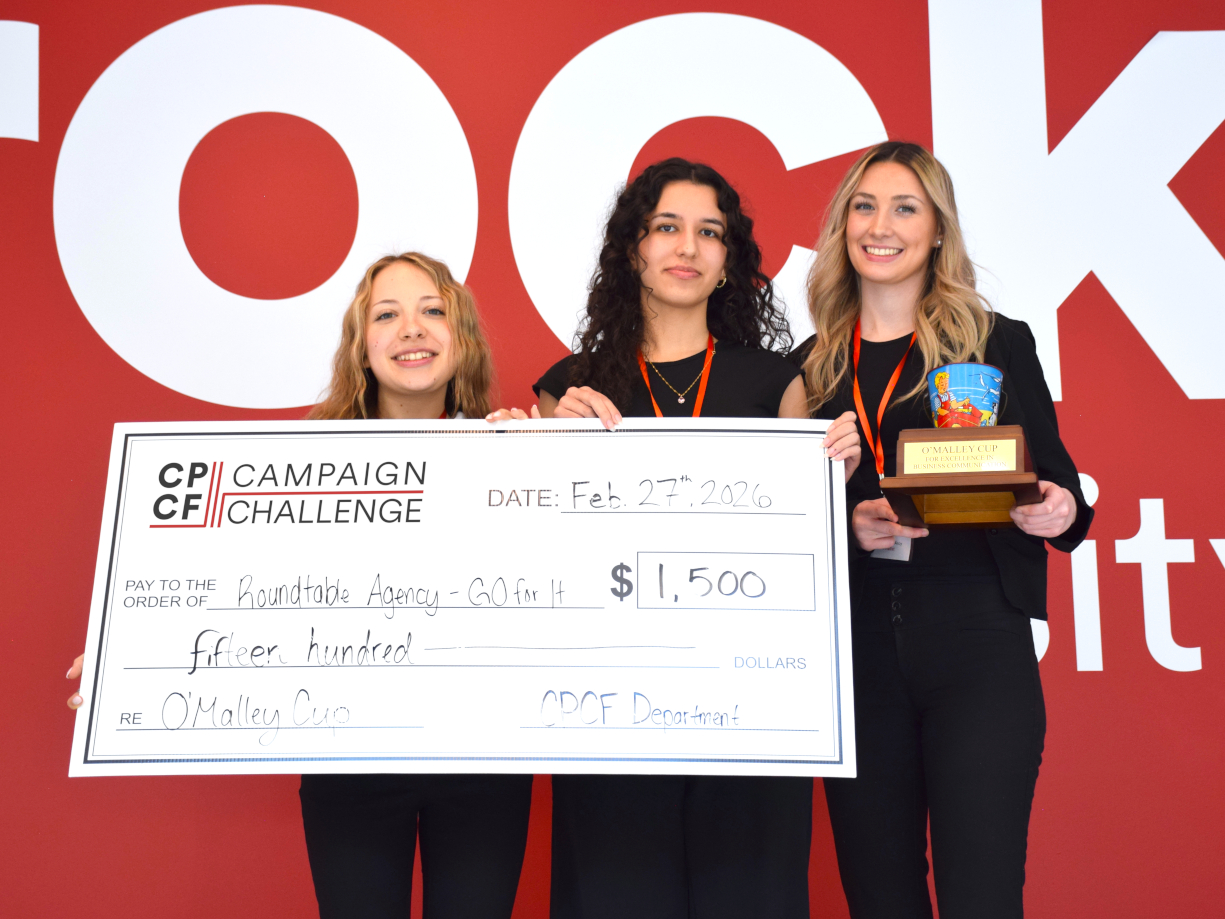 Three students pose for a photo while holding a oversized check and a trophy. Behind them is a red wall with the Brock University logo.