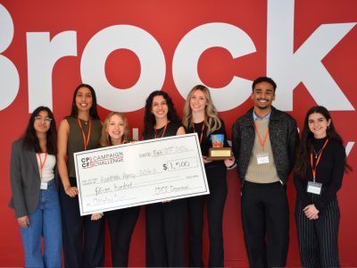 Five students and the two MCs of the event pose for a photo while holding a oversized check and a trophy. Behind them is a red wall with the Brock University logo.