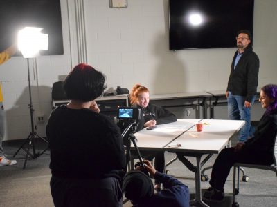 Two young women are sitting at a table preforming a scene while in the foreground a woman is recording her with camera. they are in a classroom setting. There is a man standing in the background giving direction, he is their teacher.