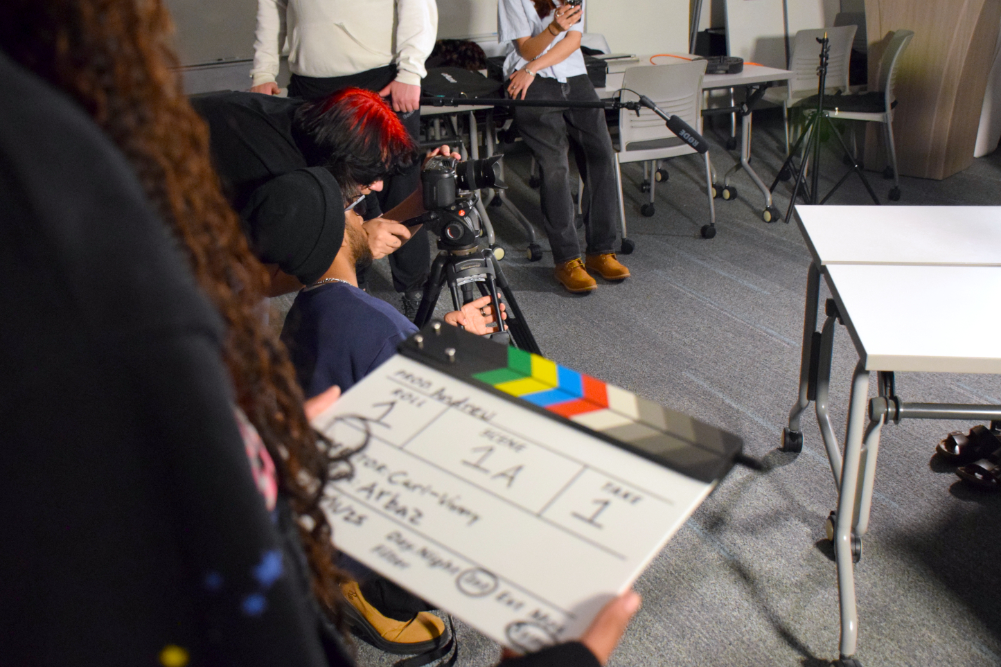 A close-up of hands holding a film slate with info on it referring to the current scene, shot and take. In the background a group of students can be seen handling production equipment, and setting up the camera.
