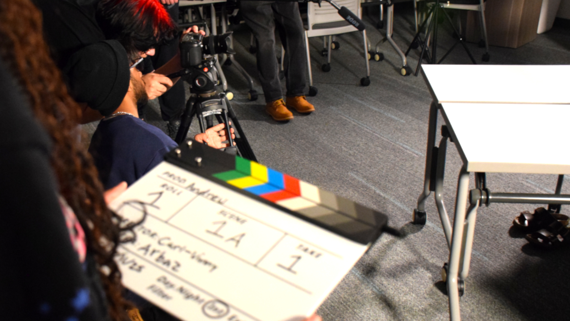A close-up of hands holding a film slate with info on it referring to the current scene, shot and take. In the background a group of students can be seen handling production equipment, and setting up the camera. 