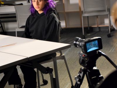 A camera in the foreground is recording an actor sitting at a table. She has purple hair and is wearing a black hoodie.