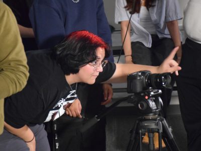 a woman with red hair crouches down to look into the camera, while motioning to another crew member with her hand.