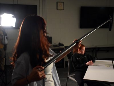 a woman with long hair is holding a boom mic out towards a performer at the table.