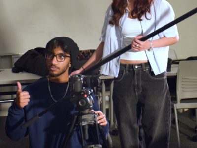 A woman with long hair hold a boom mic in-between shots, while a man crouches in front of her beside the camera an give a thumbs up in confirmation.